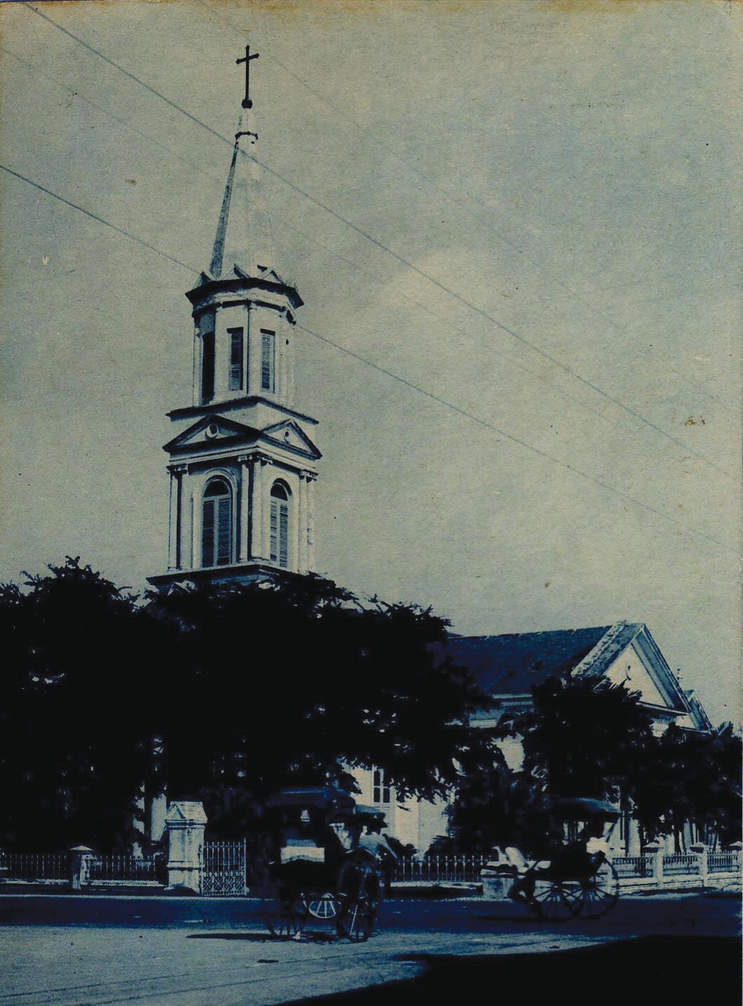 Old photograph of a church with a tall spire and cross, trees in front, and horse-drawn carriages on the street.